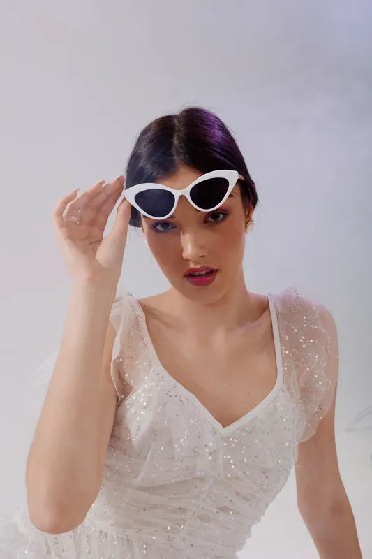 Young woman in a white sparkling dress adjusts her stylish white sunglasses. She poses confidently against a light background. Concept of woman beauty fashion and elegance.