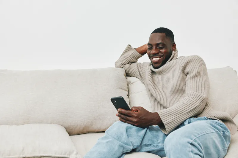 Happy young Black man relaxing on a couch, engaging with his smartphone, wearing a cozy sweater, showcasing a relaxed and joyful mood