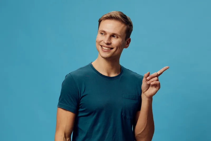 Smiling man with short hair wearing casual blue t-shirt pointing to the side on a plain blue background studio portrait. People lifestyle concept, expressing excitement and curiosity.