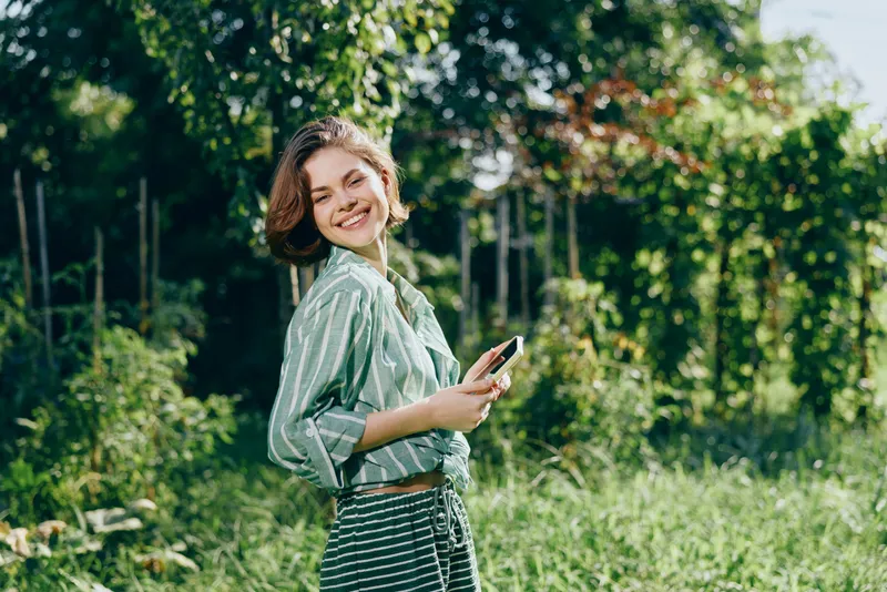A young woman stands outdoors in bright sunlight, smiling softly as she holds a smartphone amid lush green grass and trees.