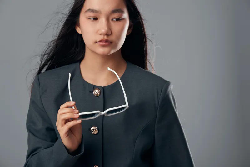 Young woman holding white eyeglasses looking down in thoughtful expression, wearing dark jacket with buttons on plain gray background in studio portrait.