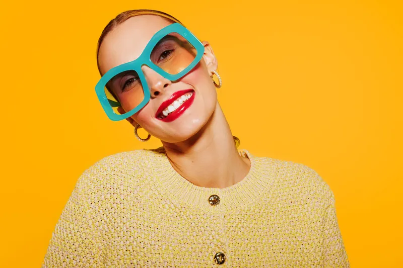 Smiling woman with bright red lipstick wears large blue sunglasses and yellow knitted sweater. Vibrant orange background creates cheerful and stylish portrait with modern fashion concept.