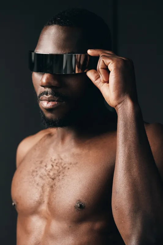 African man with dark skin wearing futuristic visor glasses in creative conceptual visual style. Portrait of shirtless male with modern technology accessory on black background.