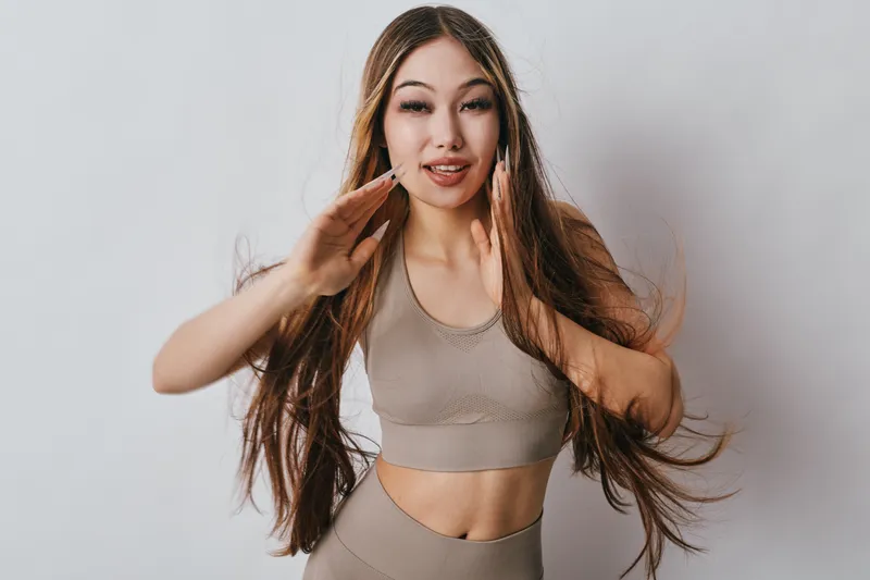 Young woman in beige sportswear posing with long hair and smiling on white background. Fitness model with playful expression in activewear for healthy lifestyle and workout.