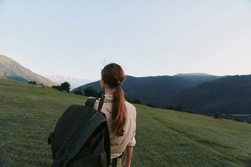 A woman with a backpack stands on a green hillside, gazing toward distant mountains. A calm, scenic moment highlighting solo outdoor exploration and simple lifestyle.