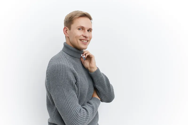 Cheerful smiling man with blond hair wearing a cozy gray turtleneck sweater pose against plain white background in a studio, conveying friendliness, warmth, positive attitude, casual style, and approa