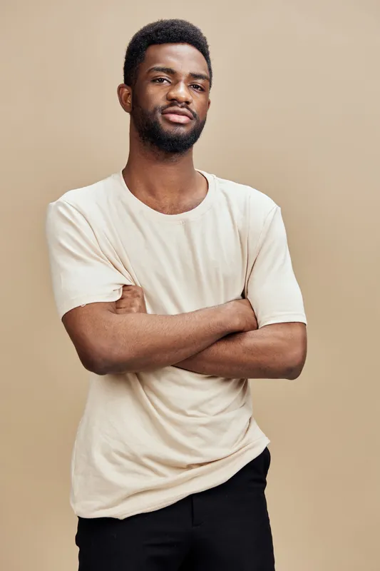 Confident young African man posing with crossed arms and a relaxed expression against a beige background, showcasing casual clothing and a modern style