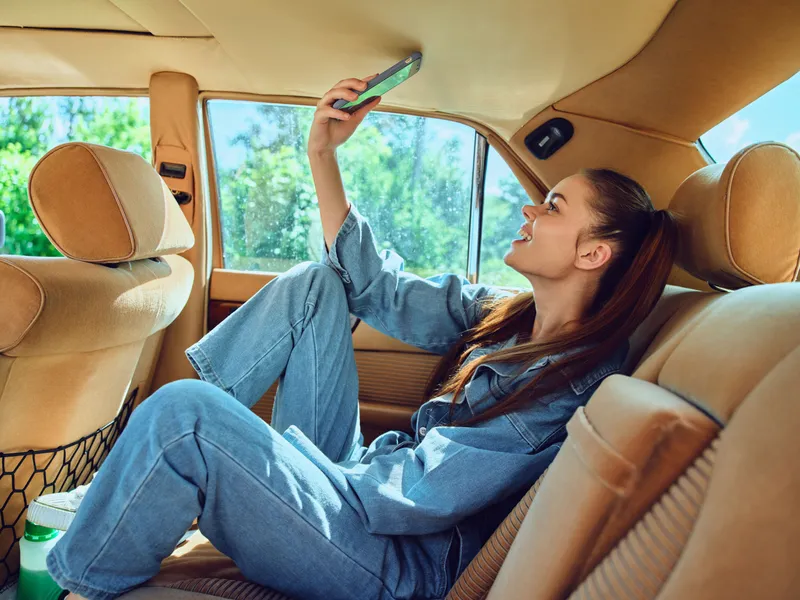 Young woman enjoying a sunny day in a vintage car, taking a selfie, wearing denim clothing, with a joyful expression and a lush green background.