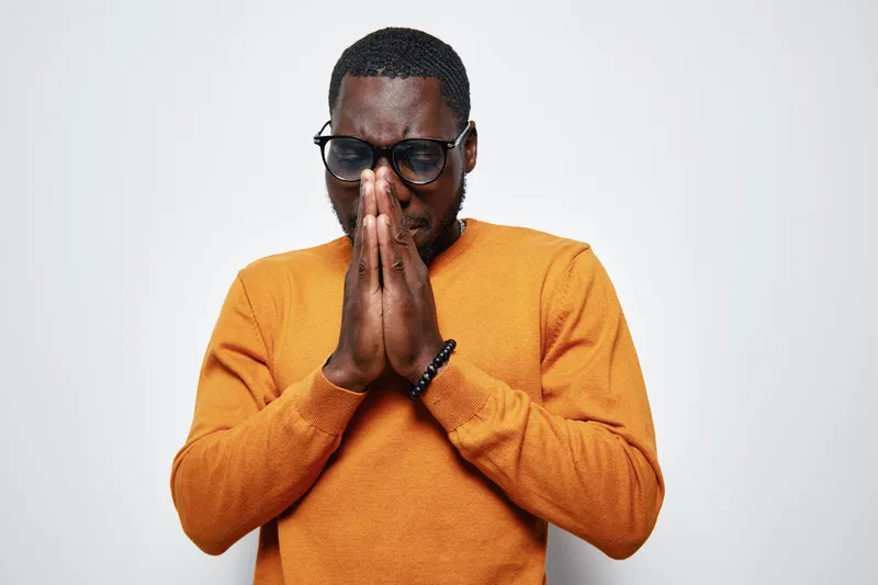 African man with dark skin and glasses wearing orange sweater praying with closed eyes and hands together isolated on white background for faith and spirituality.