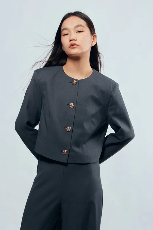 Young woman wearing a dark gray tailored jacket and matching pants posing against light background. Fashion portrait with minimalist style, long black hair and neutral expression on model face.