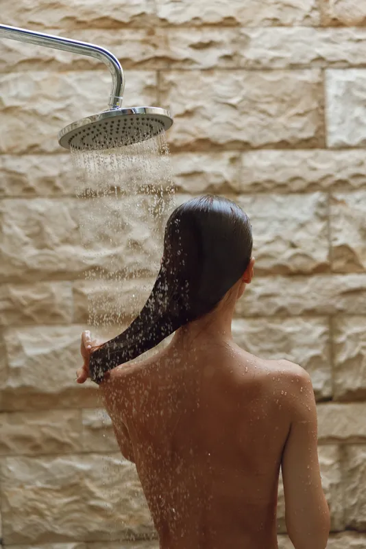 woman showering outdoors with wet hair under rainfall showerhead, natural stone wall background, calm and refreshing moment in warm lighting