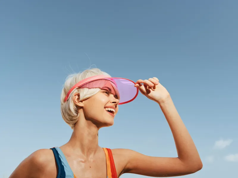 Bright portrait of a smiling woman wearing a pink visor as sunlight highlights her face against a clear blue sky, conveying a fresh summer vibe and energetic mood.