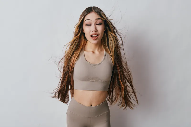 Young woman with long hair wearing beige sportswear standing with eyes closed and happy expression in studio with plain background. Fitness and wellness concept with natural look.