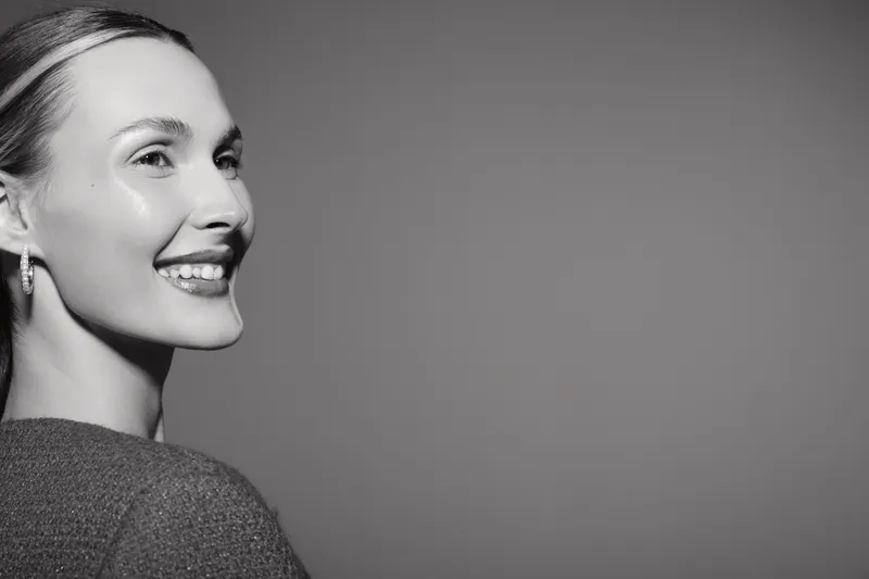 Smiling Caucasian woman with light skin and blond hair looks sideways, wearing earrings and a knitted sweater. Studio portrait with soft lighting and grey background.