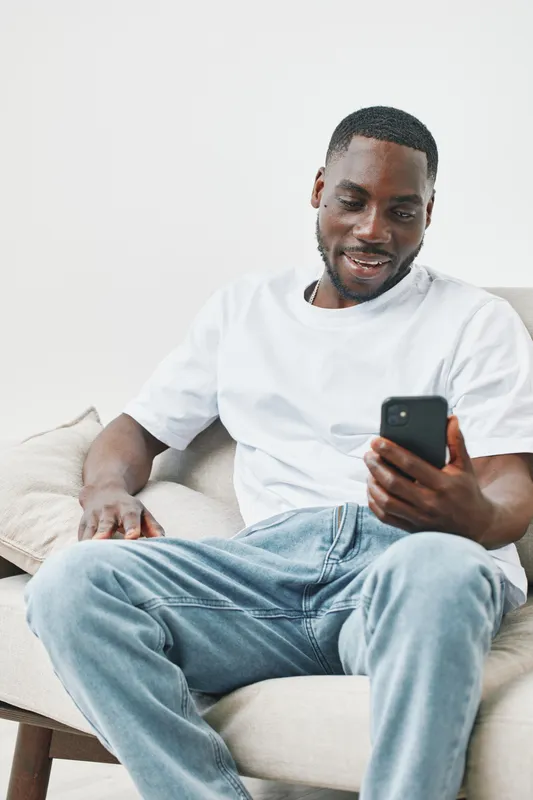 Happy young African man in casual outfit sitting on a sofa and using smartphone, enjoying social media or video chat at home