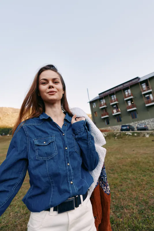 Woman smiling in denim shirt holding a jacket outdoors in a grassy field near a residential building, casual portrait with relaxed pose, natural light and confident expression.