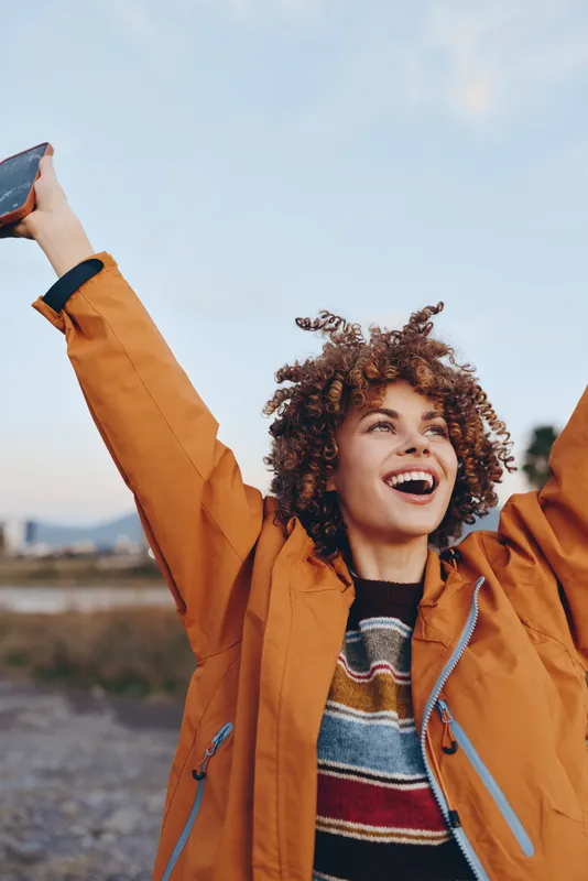 Outdoor woman lifestyle portrait by water, curly hair framing her face as she is laughing, orange jacket zipped up, one arm raised with a smartphone, bright sky and coastline