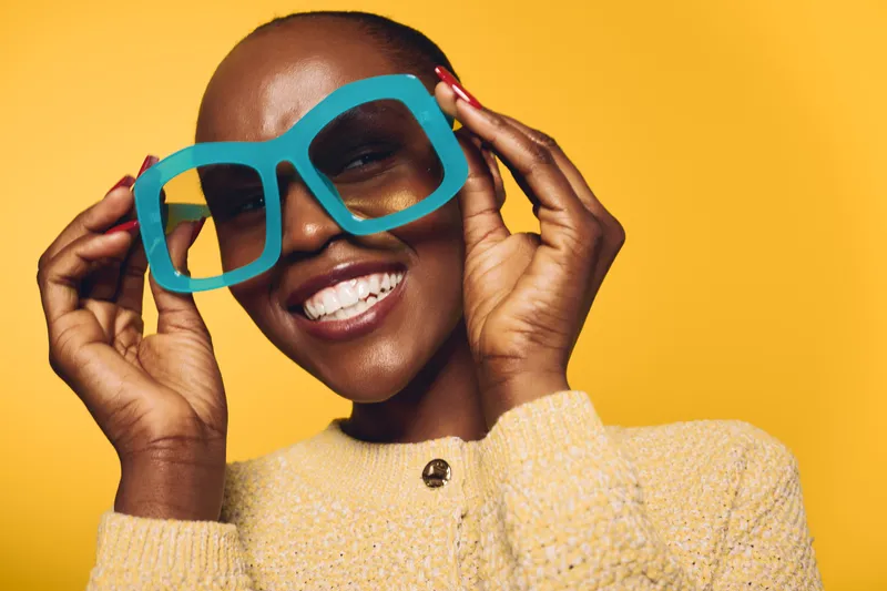 Happy African woman with dark skin wearing oversized blue glasses and a beige sweater smiles brightly against yellow background. Concept of joy, fashion, and vibrant portrait.