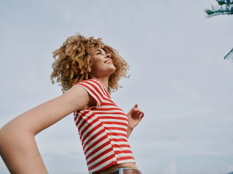 Young woman with curly hair wearing a red and white striped shirt smiles upward against a clear sky, capturing carefree youth, sunshine, and effortless outdoor style.