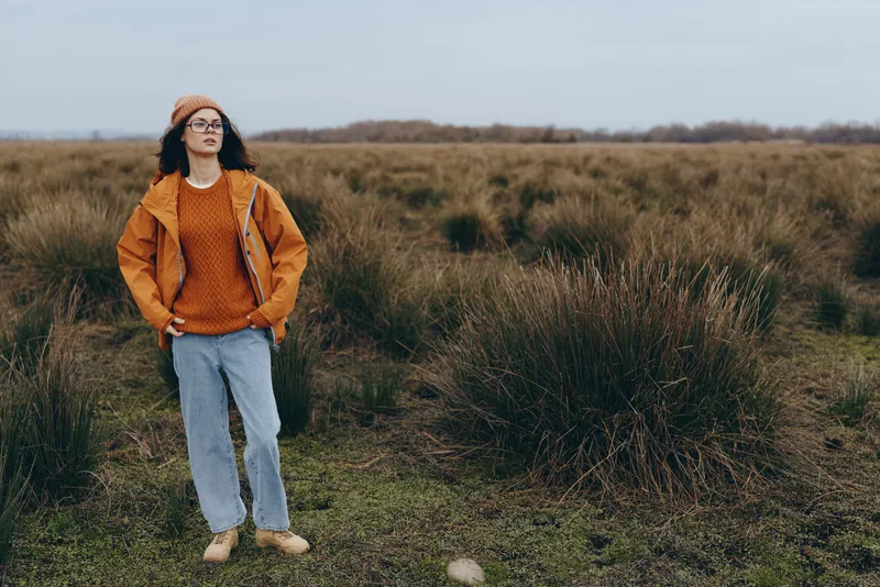 Woman lifestyle outdoors in a grassy field, orange jacket and jeans, walking along a rural path with calm expression, open landscape wide horizon nature background in soft daylight