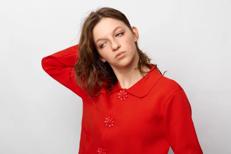 Young woman in red sweater looks away thoughtfully with hand behind head against white background. Concept of contemplation, casual fashion, and natural beauty in studio lighting.