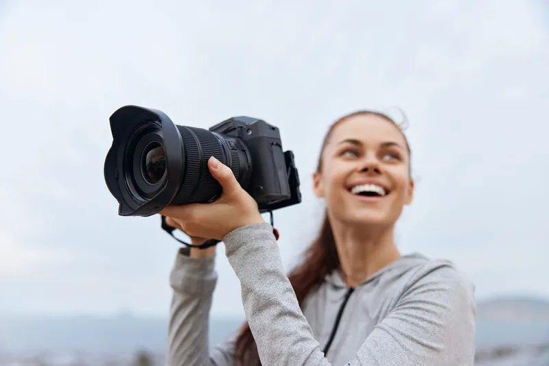 Happy young woman photographer outdoors, smiling while holding a camera, wearing a gray hoodie against a cloudy sky background, capturing memories, lifestyle concept