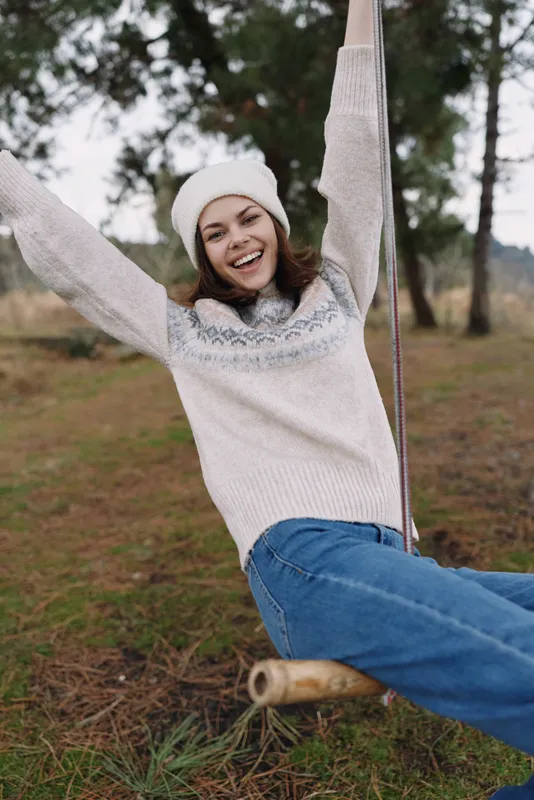 Young woman on a swing outdoors, wearing a cozy sweater and beanie, smiling joyfully in a park with trees, grass and soft daylight today