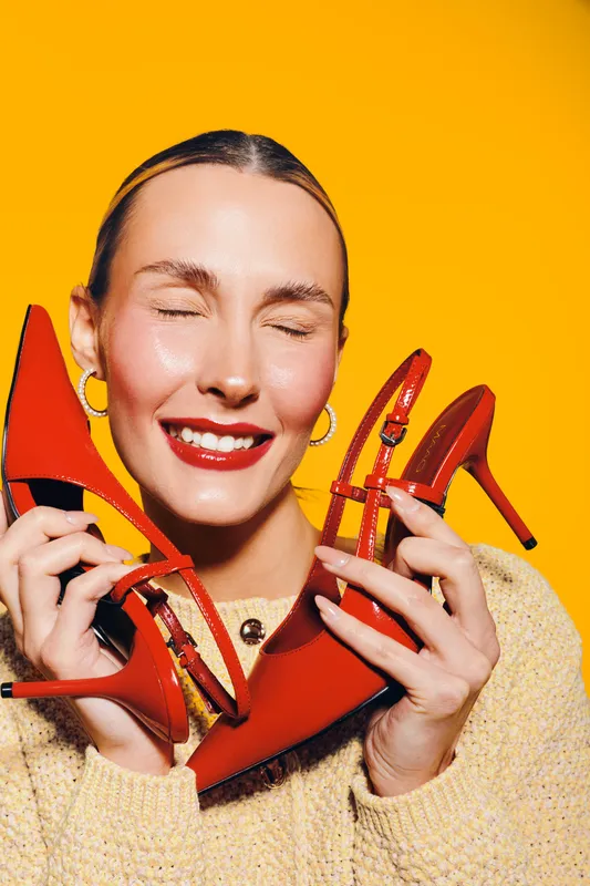 Happy woman with closed eyes holds red high heel shoes near face against yellow background. Concept of fashion, style, footwear, and joyful female expression in studio lighting.