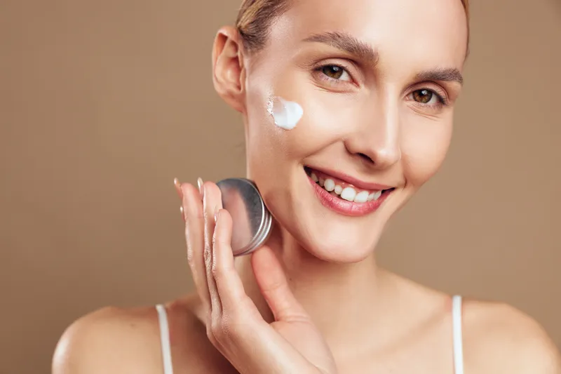 A smiling young woman applies facial cream on her cheek in a bright studio, demonstrating a casual skincare routine, clean complexion, and confident, relaxed mood.