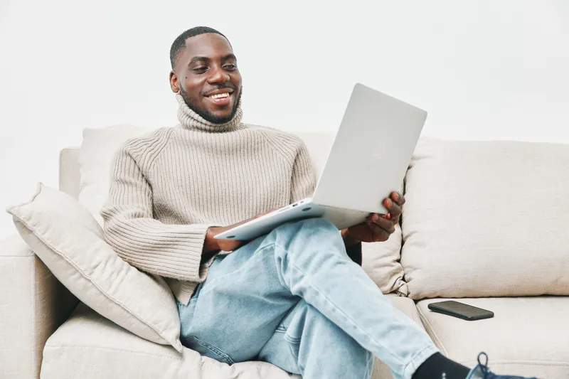Happy young man wearing a turtleneck sweater, sitting on a couch and using a laptop, showcasing modern lifestyle and remote work concept