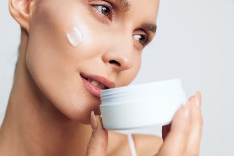 Woman skincare beauty moisturizer cream face closeup applying on cheek, woman holds a small jar of cream near lips, bright studio light, hydration selfcare concept