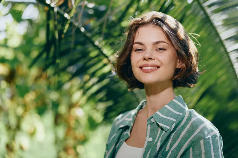 Smiling young woman outdoors in bright sunlight, wearing a green striped shirt, standing among lush palms and tropical foliage.