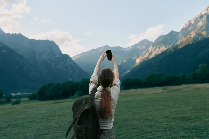 Cheerful woman outdoors, backpack on shoulder, capturing landscape with a camera against mountains, meadow and nature, conveying travel lifestyle in bright natural light