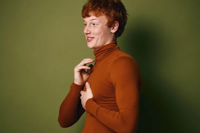 Young man with red hair wearing brown turtleneck sweater standing against a green background, smiling and adjusting collar in casual pose for fashion or lifestyle concept.