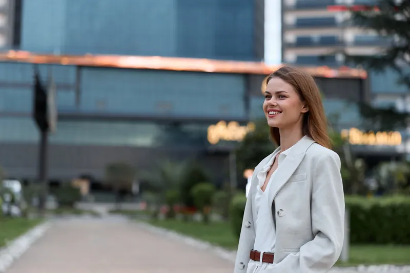 Confident woman in business attire smiling outdoors near modern architecture, exuding professionalism and empowerment during a sunny day.