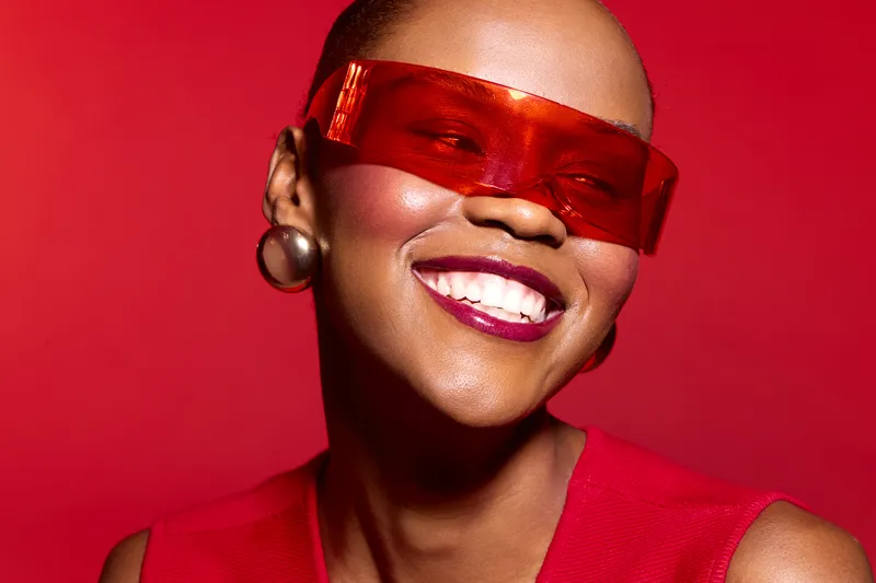 Smiling African woman with dark skin wearing red sunglasses and red top poses against red background. Concept for fashion, beauty, confidence, and modern style portrait.