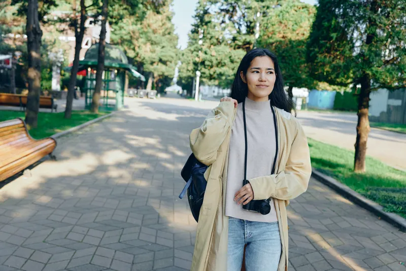 Woman walking in a park during daytime, wearing a beige coat over a sweater and jeans, carrying a backpack, showcasing casual street style amid green trees and sunlit pavement