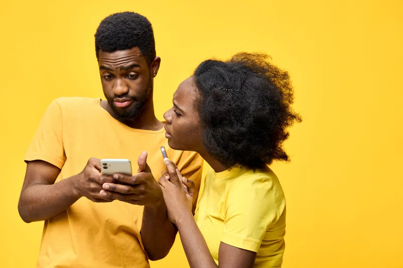 Young diverse couple using smartphones, expressing curiosity and surprise, bright yellow background, modern style, playful interaction, technology lifestyle