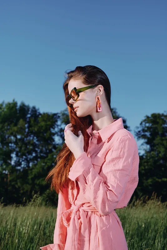 Stylish woman in a pink dress and sunglasses poses gracefully in a sunny green field, exuding confidence and serenity, ideal for fashion and lifestyle themes