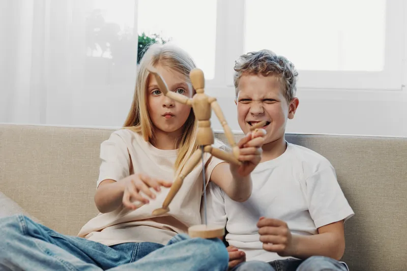Two children sitting on a sofa play with a wooden artist mannequin, the girl focused and the boy making a playful face. Concept of childhood fun, creativity, and sibling interaction.