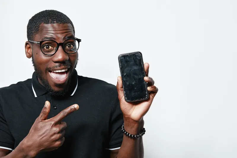 African American man with dark skin and glasses smiling and pointing at smartphone screen in black polo shirt on white background. Positive expression and casual style.