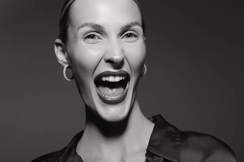 Young white woman with light skin laughing loudly in black silk blouse. Studio portrait with dramatic lighting on dark background. Concept of joy, emotion, and female expression.
