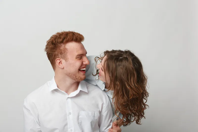 Happy young couple with red hair and curly brown hair laughing together on white background. Concept of love, joy, relationship, and positive emotions between man and woman.