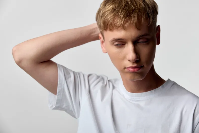 Young man in a white tee touches the back of his neck with a tired expression, conveying stress and fatigue in a minimal studio setting, calm mood and casual wear.