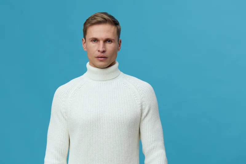 Portrait of a serious young man with fair hair and fair skin wearing a white turtleneck sweater against a plain blue background. Studio shot for fashion, portrait, or lifestyle projects.