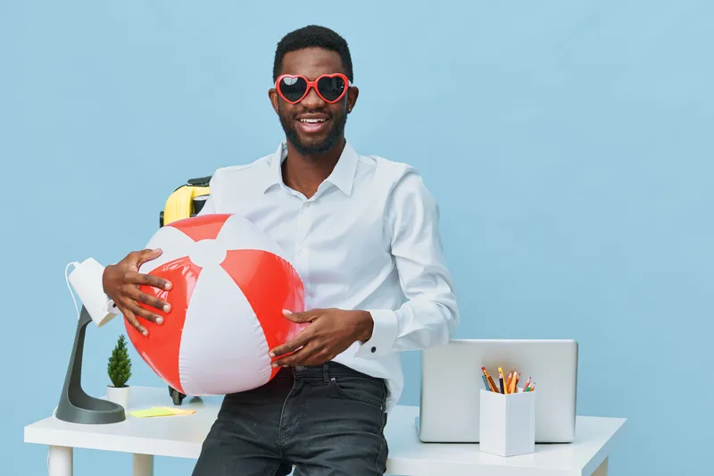Joyful young man with dark skin in white shirt and heart-shaped sunglasses holding a beach ball in a modern office space with summer accessories, creating a lively and cheerful vibe. People lifestyle 