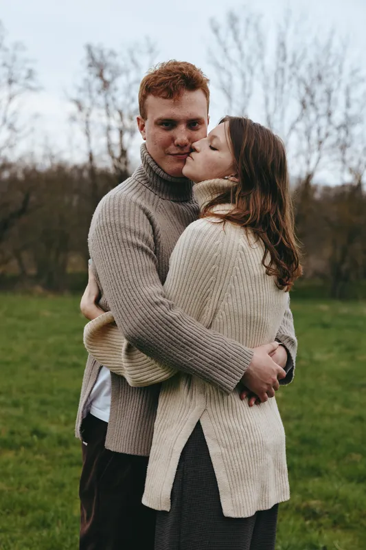 Couple in cozy sweaters share a warm embrace outdoors, capturing a candid, romantic moment in a park setting. A lifestyle portrait focused on connection, comfort, and affection between partners.