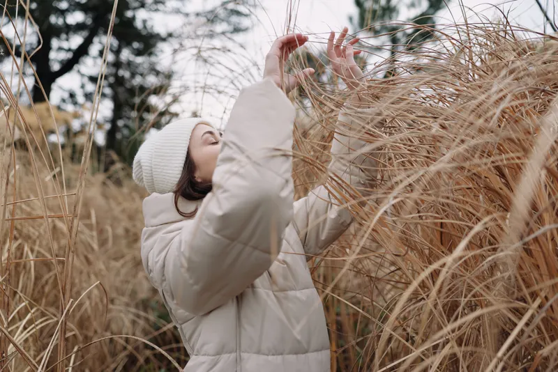 Child in a warm white jacket and knit hat reaches for tall grasses in an open field, exploring nature in winter light with curiosity and gentle motion.