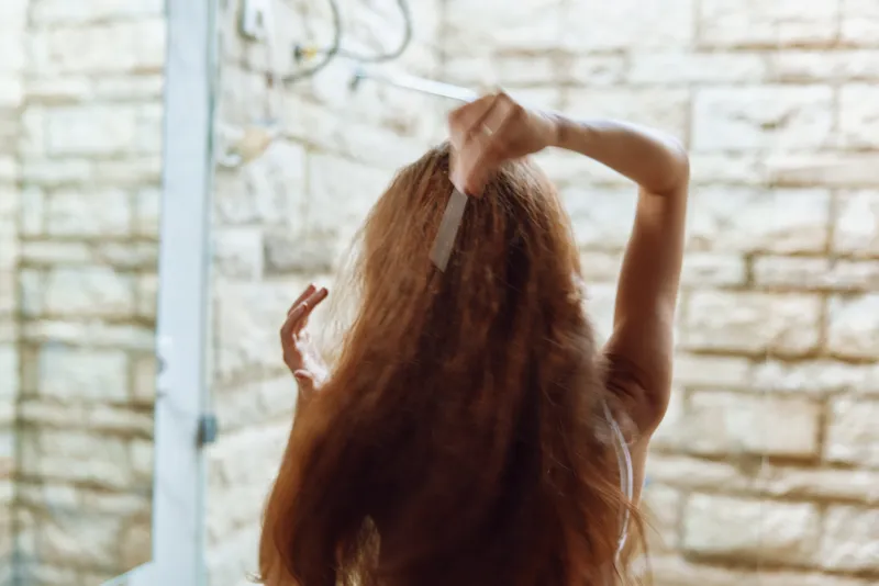 woman combing long curly hair in bathroom with white tile background, natural lighting, casual morning routine, healthy hair care
