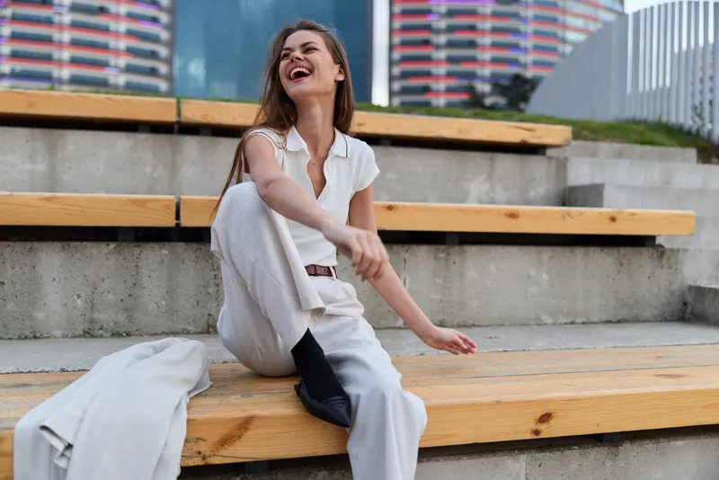 Confident businesswoman enjoying her success while sitting outdoors, dressed in a chic white outfit, exuding joy and positivity in a vibrant urban setting.